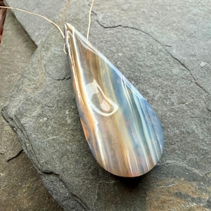 May include: A teardrop-shaped pendant with a gold wire attached. The pendant has a blend of brown, blue, and white colours, with a glossy finish. The stone is resting on a grey, textured surface.