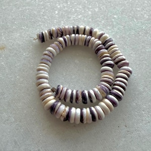 May include: A strand of disc-shaped beads in shades of white and purple. The beads are strung together, forming a circular shape. The beads appear to be made of a natural material, possibly shell or stone. The overall aesthetic is organic and earthy.