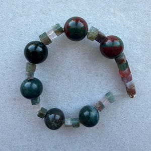 Könnte beinhalten: Ein Armband aus runden und quadratischen Edelsteinen in Grün-, Rot- und Rosatönen. Das Armband hat eine Vielzahl von Texturen und Farben und einen goldfarbenen Verschluss. Das Armband ist ein Schmuckstück.