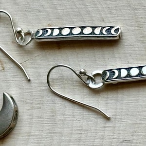 May include: A pair of silver earrings with a moon phase design. Each earring features a thin silver bar with eight moon phases, and a silver hook.