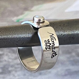 May include: A silver ring with a silver band and a silver top. The band is engraved with the word "Namaste" and a lotus flower. The top of the ring has a clear stone.
