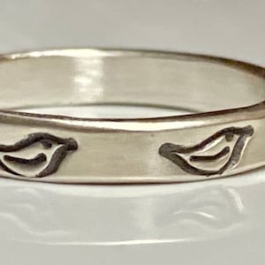 May include: A silver band ring with a textured surface and a repeating design of small birds.