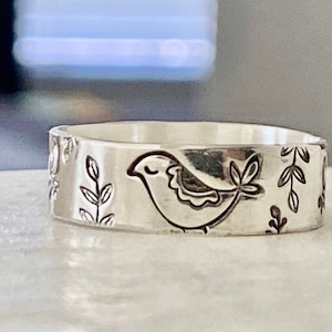 May include: A silver ring with a bird and floral design. The bird is facing right and has a closed eye. The floral design is on both sides of the bird.