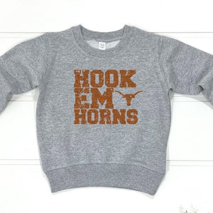 Könnte beinhalten: Ein graues Sweatshirt mit langen Ärmeln. Auf der Vorderseite des Sweatshirts steht der Text "HOOK 'EM HORNS" in einer verblassten orangefarbenen Schrift, zusammen mit einer Longhorn-Silhouette. Das Sweatshirt wird auf einer weißen Holzoberfläche präsentiert.