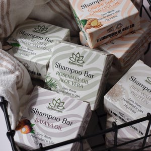 May include: A black wire basket filled with six different shampoo bars. Each bar is wrapped in white paper with a pink and white striped design. The bars are labeled with the brand name "Relaxcation" and the ingredients, such as "Ginger Root Camelina Oil", "Shea Butter Colloidal Oatmeal", "Rosemary Oil Aloe Vera", "Rice Water Protein", and "Batana Oil". Each bar is labeled "Zero Waste" and "Handcrafted in USA".