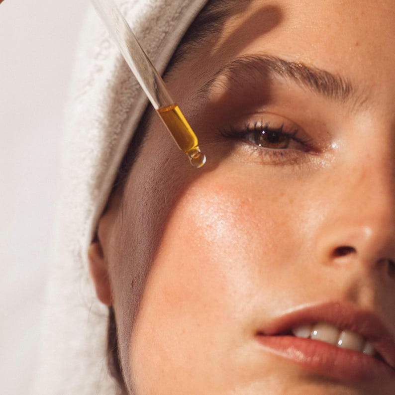Peut inclure: Gros plan sur un visage avec une serviette blanche sur la t&ecirc;te. Une pipette en verre d&eacute;pose un liquide dor&eacute; sur la peau, une goutte pend au bout. La peau semble saine et &eacute;clatante, sugg&eacute;rant un produit de soin.