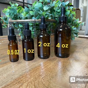 May include: Four amber glass bottles with black droppers and labels indicating their size in ounces: 0.5 oz, 1 oz, 2 oz, and 4 oz. The bottles are arranged in order of size from left to right on a wooden surface.