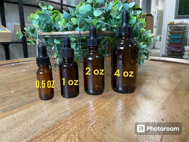 May include: Four amber glass bottles with black droppers and labels indicating their size in ounces: 0.5 oz, 1 oz, 2 oz, and 4 oz. The bottles are arranged in order of size from left to right on a wooden surface.