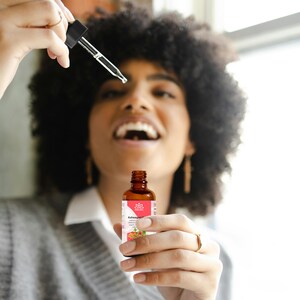 May include: A brown glass bottle with a dropper, labeled "Ashwagandha" and a pink and white lotus flower logo. The bottle is being held by a person with curly hair.