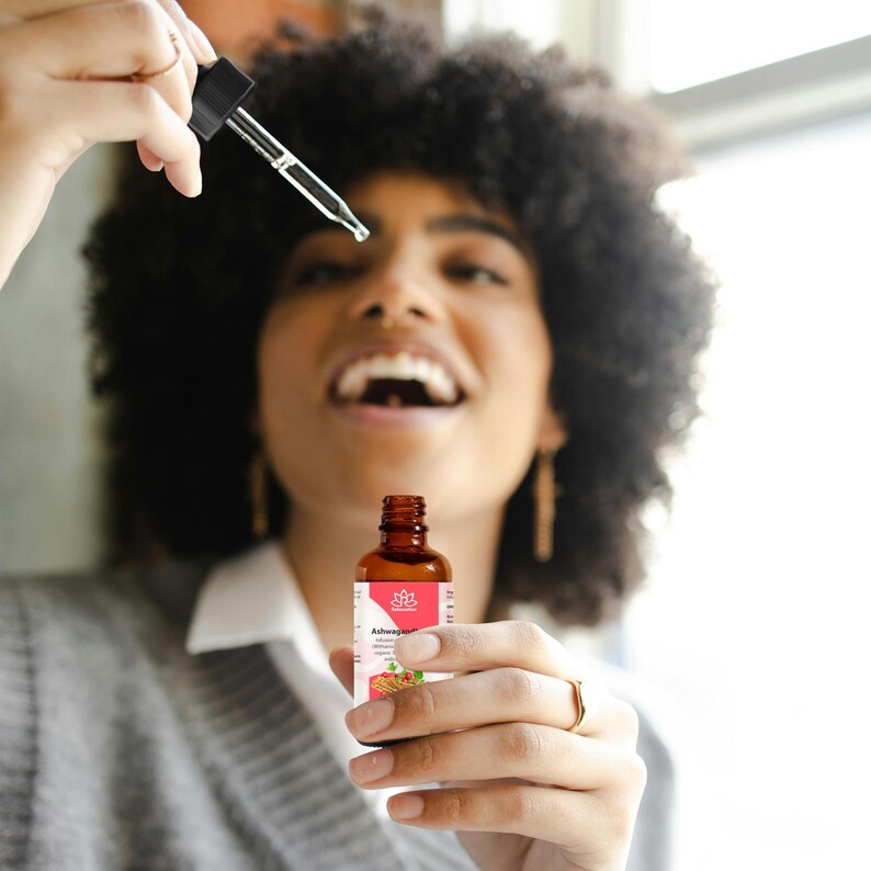 May include: A brown glass bottle with a dropper, labeled "Ashwagandha" and a pink and white lotus flower logo. The bottle is being held by a person with curly hair.