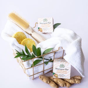 May include: Two bars of shampoo in white boxes with green leaves and ginger root illustrations. The boxes are labeled "Shampoo Bar Ginger Root Camelina Oil Zero Waste Handcrafted in USA 71g/2.5oz". A white towel, a wooden hair brush, and two round bars of shampoo are in a gold wire basket. Ginger root is on the bottom of the image.