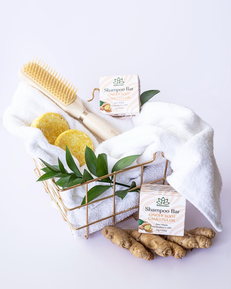 May include: Two bars of shampoo in white boxes with green leaves and ginger root illustrations. The boxes are labeled "Shampoo Bar Ginger Root Camelina Oil Zero Waste Handcrafted in USA 71g/2.5oz". A white towel, a wooden hair brush, and two round bars of shampoo are in a gold wire basket. Ginger root is on the bottom of the image.