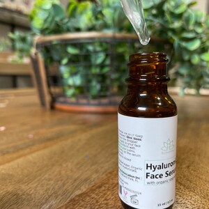 May include: A brown glass bottle with a dropper filled with a clear liquid. The bottle is labeled "Hyaluronic Face Serum with organic rose oil" and "15 ml (0.5 fl oz)".