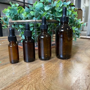 May include: Four amber glass bottles with black dropper tops of varying sizes. The bottles are arranged in a row on a wooden surface.