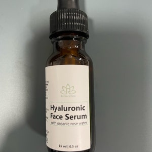 May include: A brown glass bottle with a black dropper top. The bottle is labeled "Hyaluronic Face Serum with organic rose water" and "15 ml | 0.5 oz".