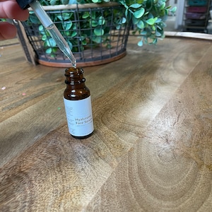 May include: A brown glass bottle with a dropper filled with clear liquid. The bottle is labeled "Hyaluronic Face Serum" with white text on a white background. The bottle is sitting on a wooden surface.