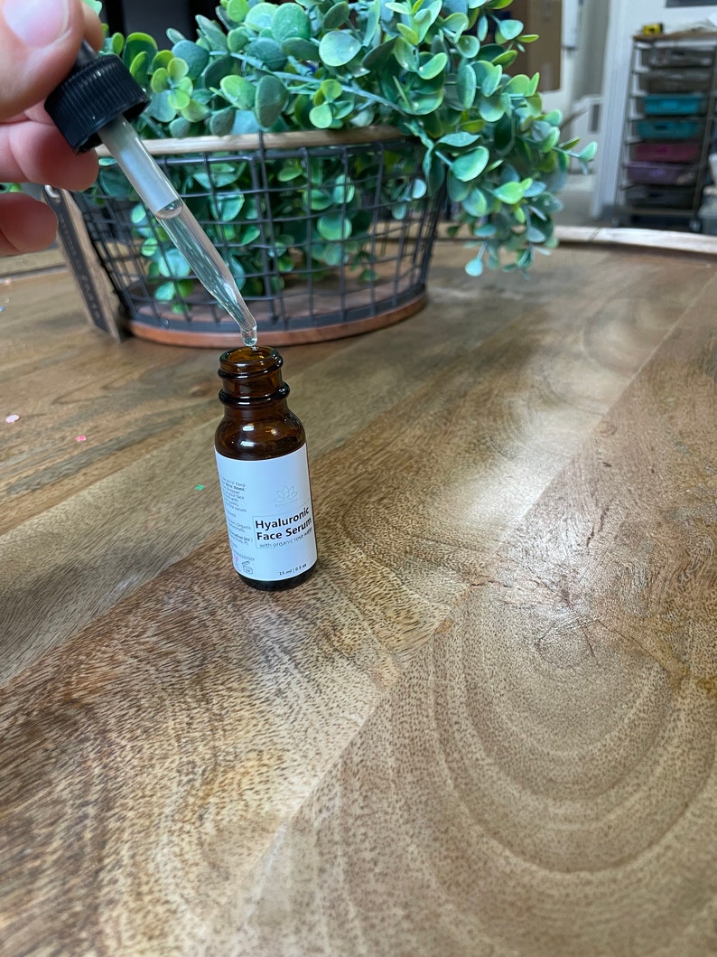 May include: A brown glass bottle with a dropper filled with clear liquid. The bottle is labeled "Hyaluronic Face Serum" with white text on a white background. The bottle is sitting on a wooden surface.
