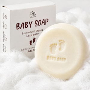 May include: A round, unscented baby soap with a footprint design, next to a white box. The box reads "BABY SOAP" and "Enriched with Organic Cocoa Butter." The soap is vegan, organic, cruelty-free, and non-GMO.