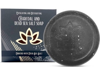May include: A black charcoal and Dead Sea salt soap bar with a white lotus flower logo. The soap is in a white box with black text that reads "Exfoliating and Detoxifying Charcoal and Dead Sea Salt Soap" and "Enriched with Dead Sea Salt".