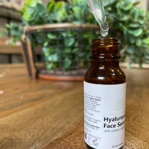 May include: A brown glass bottle with a white label that reads "Hyaluronic Face Serum with organic rosehip oil" and a dropper with a clear liquid. The bottle is sitting on a wooden surface.