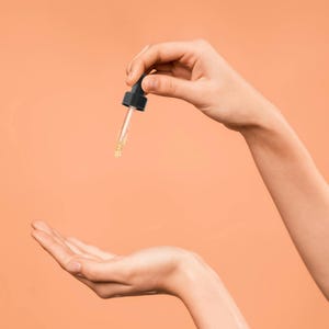 Pu&ograve; includere: Una mano tiene un flacone contagocce in vetro trasparente con un tappo nero, che rilascia un liquido trasparente su un palmo.