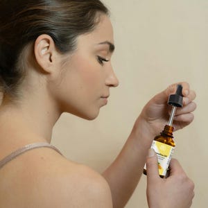 Peut inclure: Une femme tient une bouteille en verre marron avec un compte-gouttes. La bouteille a une &eacute;tiquette blanche avec un motif floral jaune et les mots "Camelina Oil". Le compte-gouttes est noir et l'huile est claire. Le fond est de couleur beige uni.