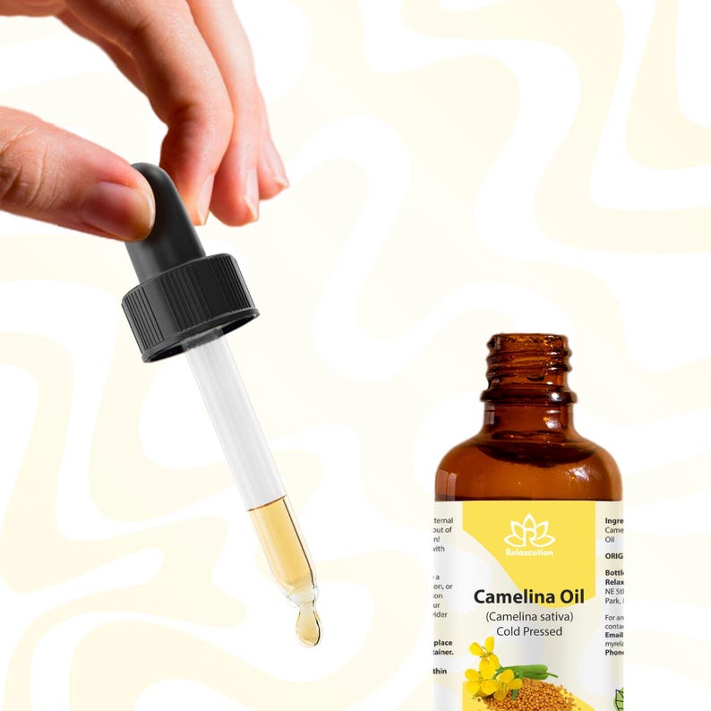 Peut inclure: Une bouteille en verre marron d'huile de cam&eacute;line avec une pipette. La pipette est tenue par une main et est remplie d'huile. La bouteille porte une &eacute;tiquette avec du texte, dont "Camelina Oil (Camelina sativa) Cold Pressed".