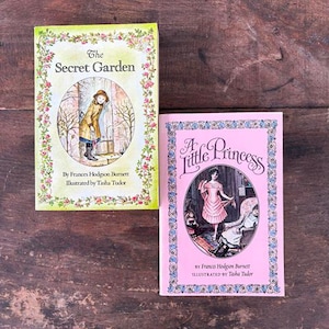 May include: Two vintage children's books, "The Secret Garden" and "A Little Princess", are displayed on a wooden surface. The Secret Garden has a green cover with floral accents and a circular illustration. A Little Princess has a pink cover with a floral border.
