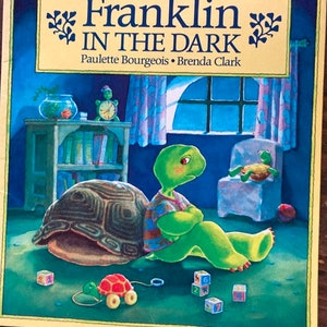 K&ouml;nnte beinhalten: Ein Kinderbuch mit dem Titel "Franklin in der Dunkelheit" von Paulette Bourgeois und Brenda Clark. Das Cover zeigt eine gr&uuml;ne Schildkr&ouml;te mit brauner Schale, die auf einem gr&uuml;nen Boden sitzt. Die Schildkr&ouml;te tr&auml;gt ein blau-wei&szlig; gestreiftes Hemd und hat einen traurigen Gesichtsausdruck. Auf dem Boden liegen bunte Bl&ouml;cke verstreut.