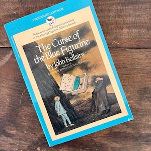 May include: A vintage paperback book titled "The Curse of the Blue Figurine" by John Bellairs. The cover features an illustration of a boy, a dark figure, and a blue figurine. The book has a blue border and is set against a wooden background.