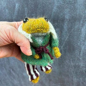 May include: A handmade knitted frog doll with a yellow head, black eyes, and a green robe tied with a burgundy belt. The frog wears white and burgundy striped pants and yellow feet. The doll is held in a hand against a dark gray background.