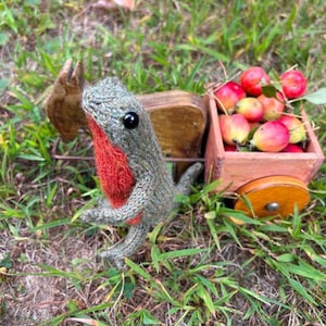 May include: A handmade knitted bird with a red breast and gray body sits next to a wooden cart filled with red and yellow apples. The cart is pulled by a wooden donkey. The scene is set in green grass.