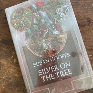 May include: A book cover with a brown background and a circular image of a person in a dark cloak riding a horse. The title of the book is "Susan Cooper Silver on the Tree".