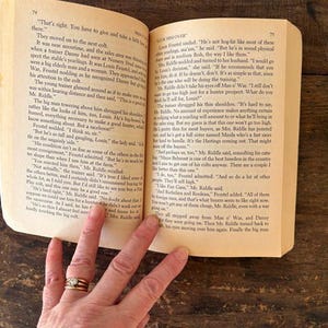 以下が含まれることがあります： 金色の指輪をした手で持たれた、テキストが書かれた開いた本。ページは黄ばんでおり、テキストは黒色です。本は木製の表面にあります。