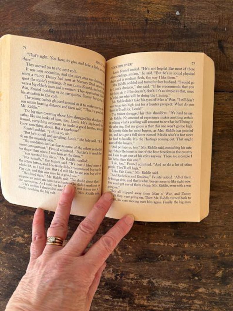 以下が含まれることがあります： 金色の指輪をした手で持たれた、テキストが書かれた開いた本。ページは黄ばんでおり、テキストは黒色です。本は木製の表面にあります。