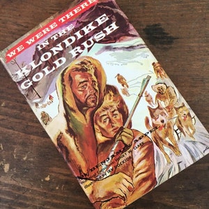 May include: A vintage book cover with a colorful illustration of people in winter clothing traveling through a snowy landscape with dogs pulling a sled. The book title is "We Were There in the Klondike Gold Rush".