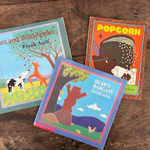 May include: Three vintage children's books with colorful illustrations. Titles include "Oats and Wild Apples," "Bear's Bargain," and "Popcorn." The books feature images of animals and nature scenes, set against a rustic wooden background.
