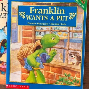 K&ouml;nnte beinhalten: Ein blaues Kinderbuch mit dem Titel "Franklin m&ouml;chte ein Haustier" von Paulette Bourgeois und Brenda Clark. Der Umschlag zeigt eine gr&uuml;ne Schildkr&ouml;te, die einen blau-wei&szlig;en Welpen h&auml;lt. Die Schildkr&ouml;te schaut in einen K&auml;fig mit einem kleinen Hund und V&ouml;geln darin. Das Buch wird von Scholastic herausgegeben.