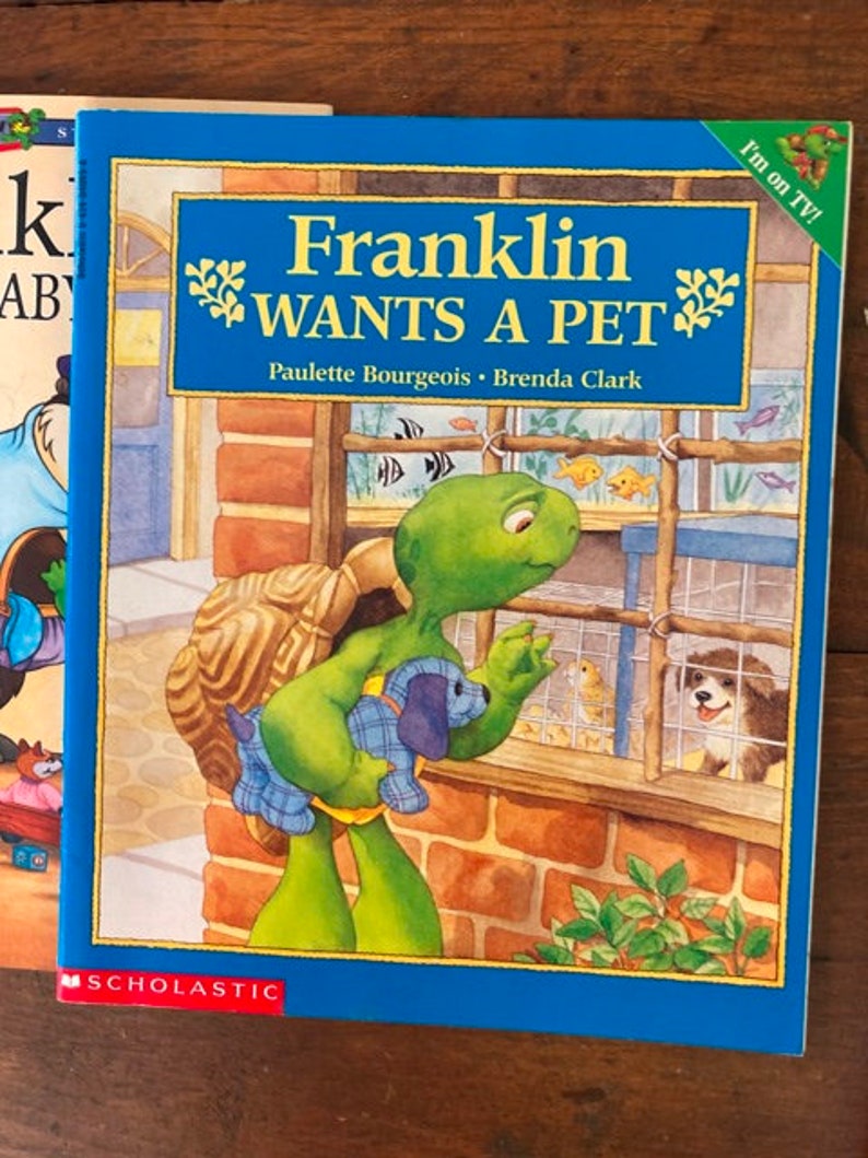 K&ouml;nnte beinhalten: Ein blaues Kinderbuch mit dem Titel "Franklin m&ouml;chte ein Haustier" von Paulette Bourgeois und Brenda Clark. Der Umschlag zeigt eine gr&uuml;ne Schildkr&ouml;te, die einen blau-wei&szlig;en Welpen h&auml;lt. Die Schildkr&ouml;te schaut in einen K&auml;fig mit einem kleinen Hund und V&ouml;geln darin. Das Buch wird von Scholastic herausgegeben.