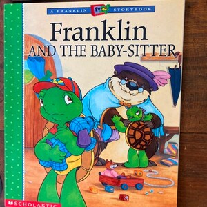 K&ouml;nnte beinhalten: Eine gr&uuml;ne Schildkr&ouml;te namens Franklin h&auml;lt eine blau-wei&szlig; gestreifte Babyschildkr&ouml;te. Die Schildkr&ouml;te tr&auml;gt einen roten Hut. Der Buchumschlag lautet "Franklin und das Kinderm&auml;dchen" und ist ein Scholastic-Buch.