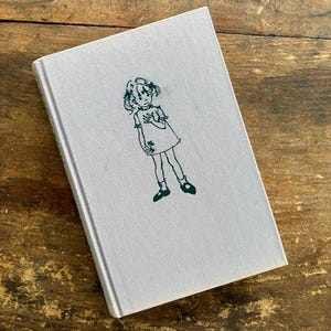 May include: A hardcover book with a light gray cover featuring a green line drawing of a girl. The drawing depicts a girl in a dress holding a flower. The book is resting on a wooden surface.