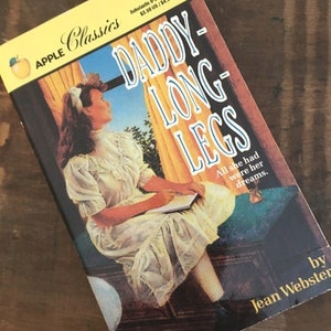 May include: A vintage paperback book titled "Daddy-Long-Legs" by Jean Webster. The cover features a woman in a white dress looking out a window. The book is part of the Apple Classics series.