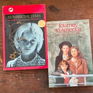 May include: Two paperback books, both with red covers. The book on the left is titled "Number the Stars" by Lois Lowry and features a girl with blonde hair. The book on the right is titled "Journey to America" by Sonia Levitin and features a group of people standing on a boat.