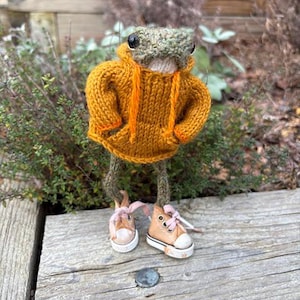May include: A handmade frog doll wearing a mustard yellow hoodie with orange drawstrings and miniature tan sneakers with pink laces. The frog's head is green and tan, and the doll is posed on a wooden surface.