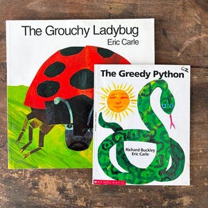 May include: Two children's books, "The Grouchy Ladybug" and "The Greedy Python," are displayed on a wooden surface. The Grouchy Ladybug features a red ladybug illustration. The Greedy Python has a green snake and yellow sun.