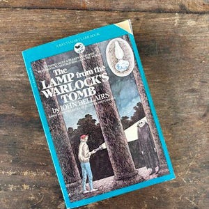 May include: A vintage paperback book titled "The Lamp from the Warlock's Tomb" by John Bellairs. The cover features an illustration of a person with a flashlight and a cloaked figure. The book has a teal and blue cover.