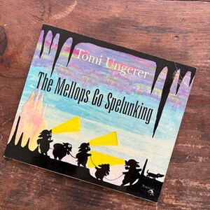 May include: A vintage children's book titled "The Mellops Go Spelunking" by Tomi Ungerer. The cover features a colorful illustration of the Mellops family with flashlights exploring a cave. The book is set against a wooden background.