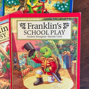 K&ouml;nnte beinhalten: Ein rotes Buch mit dem Titel "Franklin's School Play" von Paulette Bourgeois und Brenda Clark. Das Cover zeigt eine gr&uuml;ne Schildkr&ouml;te, die als Spielzeugsoldat verkleidet ist und ein Schwert und einen Hut h&auml;lt. Die Schildkr&ouml;te steht auf einer B&uuml;hne mit einem Scheinwerfer, der auf sie gerichtet ist. Im Hintergrund befinden sich Spielzeugsoldaten und ein Weihnachtsbaum.