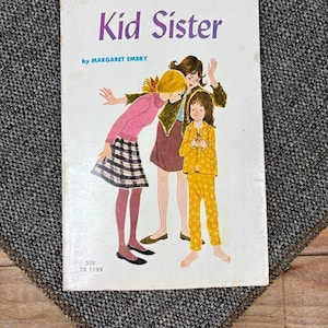 May include: Vintage paperback book titled "Kid Sister" by Margaret Embry. The cover features an illustration of three young girls in colorful outfits. The book has a white cover with purple and blue text. The book is a vintage item.