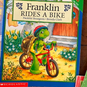 K&ouml;nnte beinhalten: Ein Kinderbuchcover mit einer gr&uuml;nen Schildkr&ouml;te, die einen Helm tr&auml;gt und ein rotes Fahrrad f&auml;hrt. Der Buchtitel lautet "Franklin Rides a Bike" von Paulette Bourgeois und Brenda Clark. Das Cover zeigt au&szlig;erdem einen blauen Himmel, gr&uuml;nes Gras und gelbe Blumen.